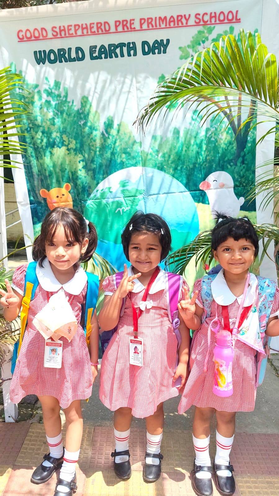 Three smiling girls in pink checked uniforms with school bags pose in front of a Good Shepherd Pre Primary School “World Earth Day” backdrop