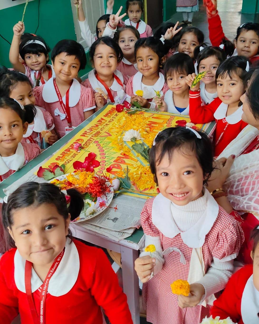 Smiling children in red and pink checked uniforms gather around a “Happy Holi” flower-petal rangoli made by the Nursery group at Good Shepherd Pre Primary School, Chandmari, Guwahati