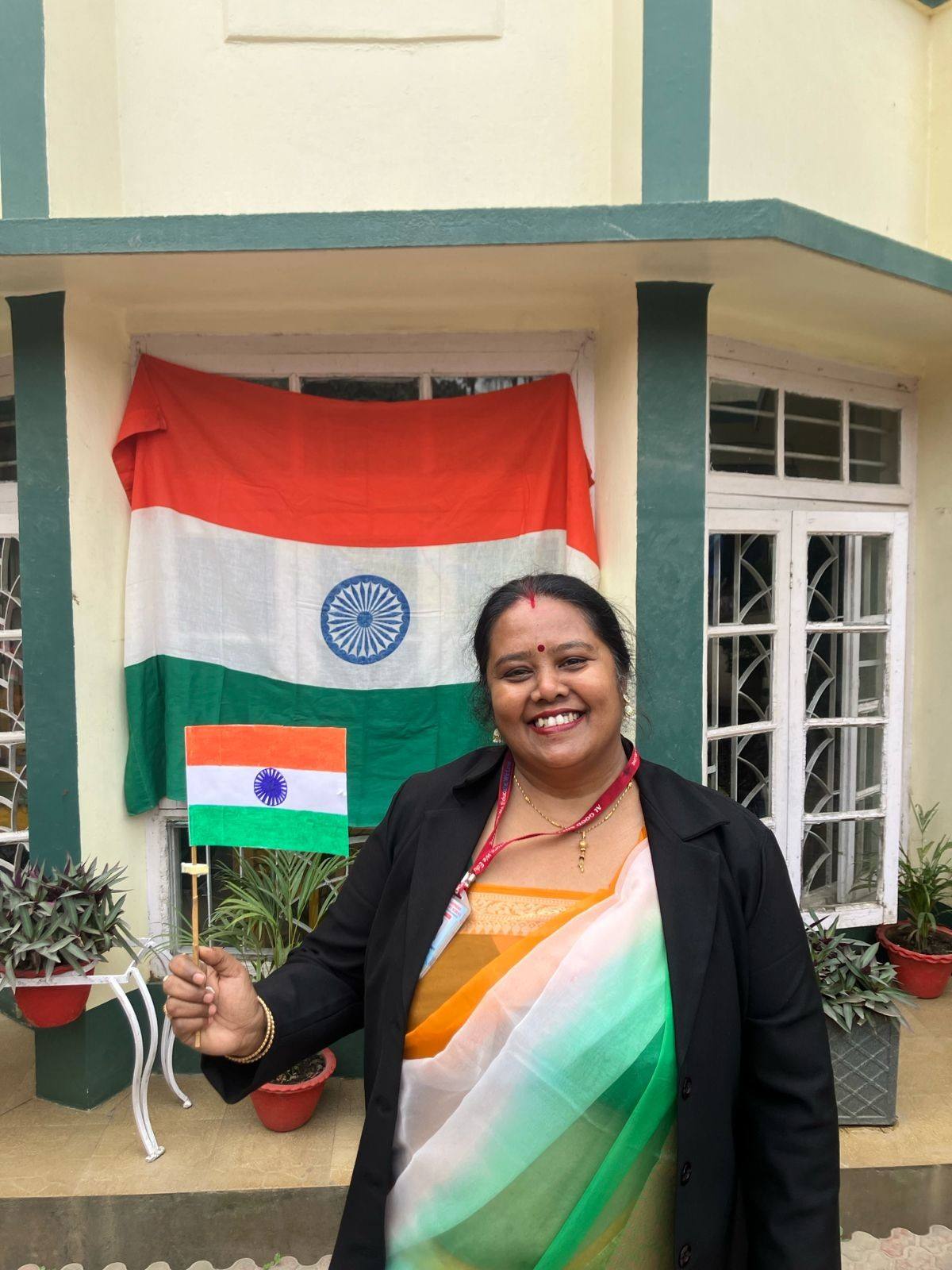 A smiling teacher in a tricolour saree holds a small Indian flag in front of the school building, with a large Indian flag hung on the window behind her